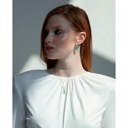 Model wearing elegant jewelry set with green stones and gold chain, highlighting a sleek hairstyle and minimalist white outfit.